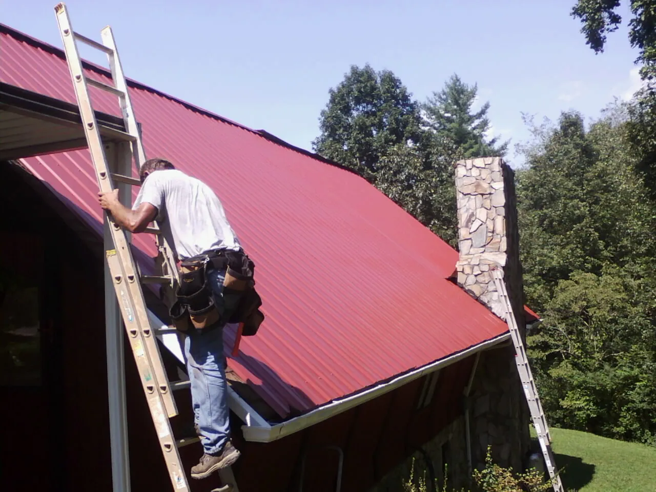 Roofer on a ladder beside a red metal roof and stone chimney — Soffit & Fascia Repair by Roofing Brothers in Pinckneyville